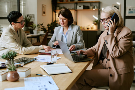Three middle aged Caucasian women collaborating at office table, one speaking on smartphone while...