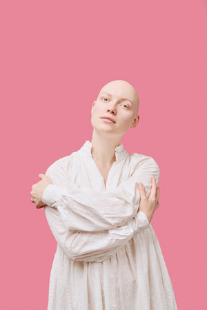 Portrait of young adult Caucasian woman with alopecia standing against pink background, crossing arms over chest, looking directly at camera with neutral expressionの写真素材