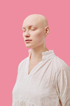 Caucasian young adult woman with alopecia standing with eyes closed against pink background, showing smooth bald scalp and serene facial expression, wearing light textured blouseの写真素材