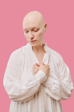 Caucasian young adult woman with alopecia standing against pink background, looking down with hands clasped near chest, showing smooth bald scalp and gentle facial expressionの写真素材