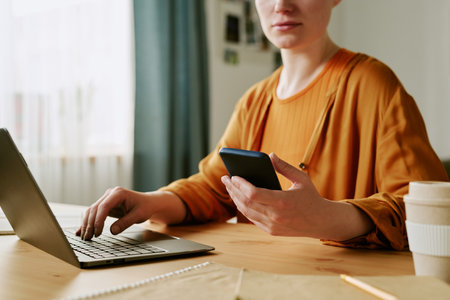 Young adult Caucasian woman sitting at desk using laptop and holding smartphone, working in home office settingの写真素材