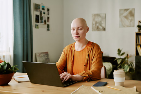 Caucasian young adult woman with alopecia sitting at desk using laptop, looking at screen with neutral expression, notebook and coffee cup on table, indoor home office settingの写真素材
