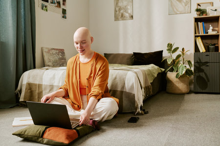 Caucasian young adult woman with alopecia sitting cross legged on floor using laptop in bedroom, smiling and engaging with device, notebook and smartphone placed nearbyの写真素材