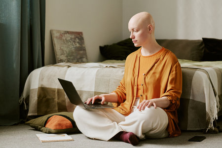 Young adult Caucasian woman with alopecia sitting cross legged on floor using laptop, looking at screen, notebook and pillow nearby, bed and wall art in backgroundの写真素材