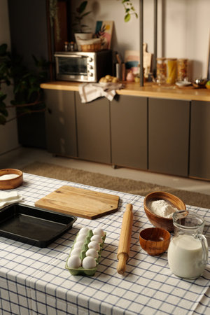 Kitchen table displaying baking ingredients including eggs, flour, milk, rolling pin, wooden bowls, baking tray and cutting board with modern kitchen background visibleの写真素材