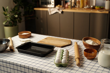 Kitchen table holding eggs in carton, rolling pin, wooden cutting board, glass jug of milk, bowls with flour and sugar, baking tray, checkered tablecloth, blurred kitchen backgroundの写真素材