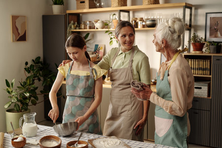 Senior woman talking with middle aged woman while teenage girl mixing ingredients in metal bowl on kitchen counter, all wearing aprons, surrounded by plantsの写真素材