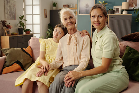 Senior woman sitting on sofa with young adult woman and child girl, all holding hands and smiling, showing multigenerational family connectionの写真素材