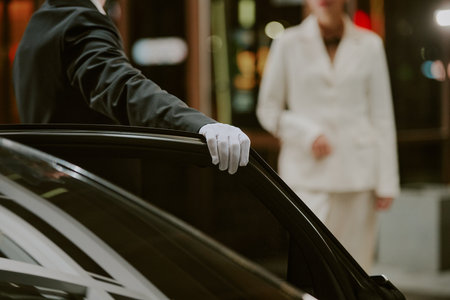 Caucasian man in suit wearing glove opening car door for Caucasian woman in business attire approaching vehicle, both appearing as middle aged professionals in urban settingの写真素材