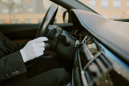 Caucasian middle aged man wearing gloves driving luxury car, hands gripping steering wheel, dashboard and interior details visible, business attire suggesting professional chauffeurの写真素材