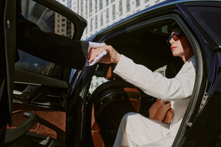 Caucasian middle aged woman exiting car while holding hand of man in suit, wearing sunglasses, business district buildings visible in background, formal interaction occurringの写真素材