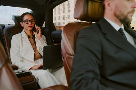 Caucasian middle aged woman sitting in back seat of car talking on phone and working on laptop while unrecognizable man driving, both wearing business attire, city buildings visible outside windowの写真素材