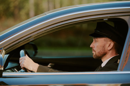 Caucasian middle aged man driving car wearing uniform and cap, sitting in driver seat with hands on steering wheel, looking forward through windshield, side profile visibleの写真素材