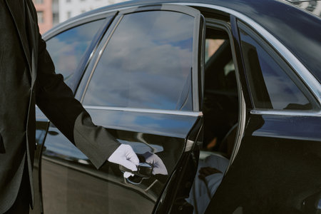 Caucasian man in formal suit and gloves opening car door, only arm and hand visible, luxury vehicle reflecting cityscape, professional chauffeur service conceptの写真素材