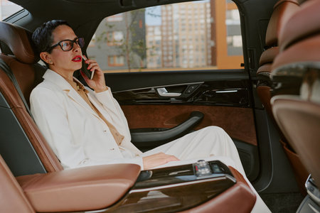 Middle aged Caucasian woman sitting in back seat of luxury car talking on smartphone, wearing glasses, short hair visible, city buildings seen through windowの写真素材