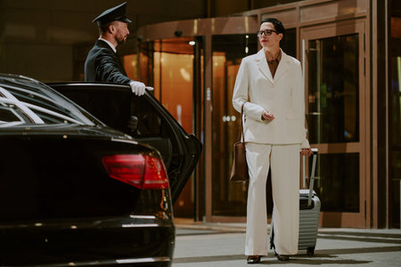 Middle aged woman standing outside hotel entrance holding suitcase and handbag while chauffeur in uniform opening car door, both engaging in business travel scenarioの写真素材