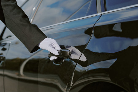 Caucasian middle aged man wearing glove opening car door, only hand and part of arm visible, luxury vehicle reflecting surroundings, professional chauffeur service conceptの写真素材