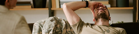 Middle aged Caucasian man wearing military uniform sitting on couch holding head in distress during...