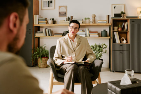 Middle aged Caucasian woman therapist sitting in office holding pen and notebook listening to unseen male client during psychological counseling session, bookshelves in backgroundの写真素材