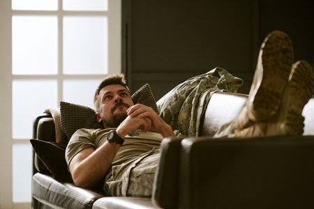 Caucasian young adult man lying on couch clasping hands together looking contemplative, wearing...