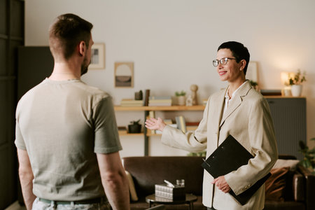 Middle aged woman therapist holding clipboard gesturing while talking to young adult man during psychological counseling session in office settingの写真素材