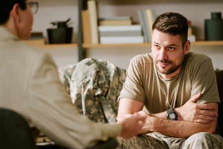 Caucasian young adult man in military uniform sitting with arms crossed engaging in therapy session with female psychologist, focusing on mental health and psychological supportの写真素材