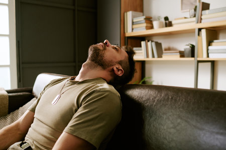 Caucasian young adult man sitting on sofa leaning head back with eyes closed appearing stressed or exhausted in home setting, illustrating psychological fatigue or burnoutの写真素材