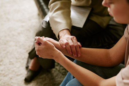 Middle aged Caucasian woman comforting young adult Caucasian woman by holding hands during therapy session, therapist supporting client in psychological counseling settingの写真素材