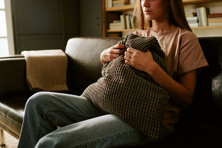 Caucasian young adult woman sitting on sofa holding textured pillow tightly against chest, showing signs of anxiety or emotional distress in home setting related to psychologyの写真素材
