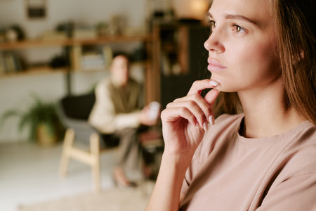 Caucasian young adult woman contemplating with hand on chin in foreground, psychologist sitting blurred in background during therapy session in modern office settingの写真素材