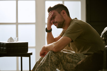 Middle aged Caucasian man sitting with hand on forehead, showing signs of emotional distress wearing military uniform in bright room suggesting psychological struggle or mental health issueの写真素材