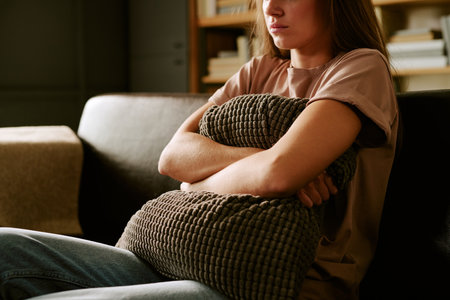 Caucasian young adult woman sitting on sofa hugging textured pillow with crossed arms, looking away with serious expression, conveying psychological distress or emotional struggleの写真素材