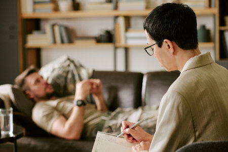 Middle aged man lying on couch talking, while middle aged woman psychologist sitting in foreground writing notes during therapy session in office settingの写真素材