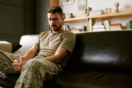 Caucasian young adult man sitting on couch wearing military uniform, looking forward with serious expression, hands clasped, conveying psychological stress or contemplationの写真素材