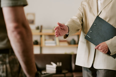 Middle aged Caucasian man extending hand for handshake toward young adult man during psychological counseling session, holding clipboard, engaging in professional interactionの写真素材