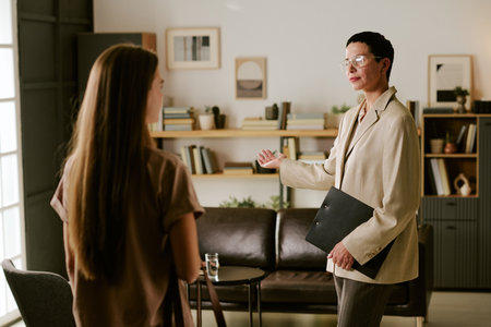 Middle aged Caucasian woman holding clipboard gesturing while talking to teenage girl in modern office setting, discussing psychological counseling or therapy sessionの写真素材