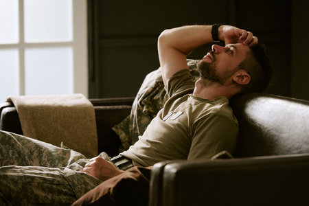 Caucasian young adult man in military uniform sitting on sofa leaning back with hand on forehead appearing stressed reflecting psychological struggle in home environmentの写真素材
