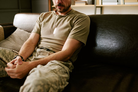 Middle aged Caucasian man sitting on couch wearing military dog tags and camouflage pants, holding hands together, showing contemplative posture in psychological contextの写真素材