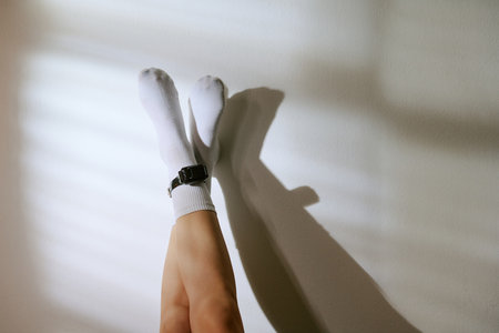 Legs of woman lying on floor with legs crossed wearing electronic ankle monitor, sunlight casting shadows on wall, concept of house arrest or electronic monitoringの写真素材