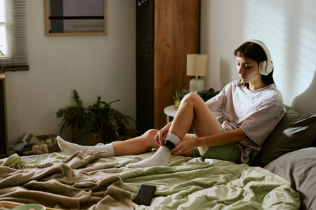 Young adult biracial woman sitting on bed wearing headphones adjusting electronic ankle monitor, staying at home with monitoring device visible on leg in domestic settingの写真素材