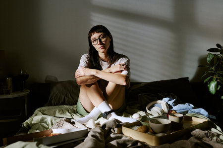 Young adult biracial woman sitting on bed with arms wrapped around knees, surrounded by food and personal items in home environmentの写真素材
