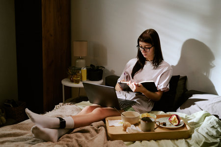 Young adult biracial woman sitting on bed working on laptop and writing in notebook with electronic ankle monitor visible on leg, breakfast tray with food and drink nearbyの写真素材