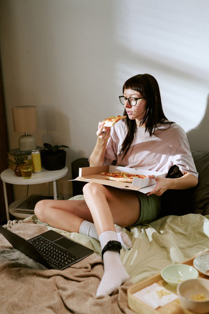 Young adult biracial woman sitting on bed eating pizza with ankle monitor visible on leg using laptop, staying at home under house arrest electronic monitoring conceptの写真素材