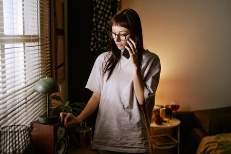Young adult biracial woman standing indoors talking on smartphone wearing ankle monitor, staying at home under supervision electronic monitoring visible in domestic settingの写真素材