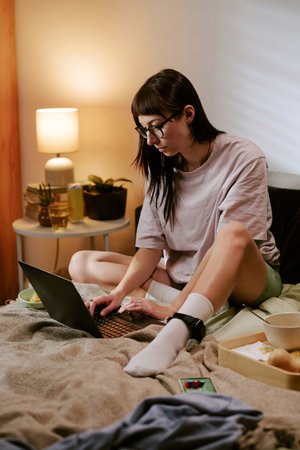 Young adult biracial woman sitting on bed using laptop with electronic ankle monitor visible on leg, staying at home and focusing on work or communication in domestic settingの写真素材
