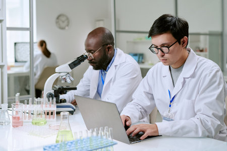 Caucasian man and Black man working in laboratory, one using laptop while other examining sample with microscope, laboratory glassware and equipment visibleの写真素材