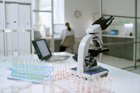 Laboratory table displaying microscope, test tubes with liquid samples, pipette and rack, background showing scientist working at computer in modern medical laboratoryの写真素材