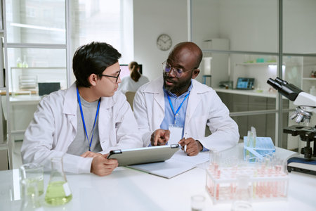 Young adult Asian man and middle aged Black man discussing laboratory results using digital tablet and notebook, sitting at laboratory table with microscope and glassware visibleの写真素材