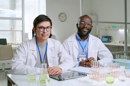 Caucasian young adult man and Black middle aged man wearing lab coats sitting at laboratory table smiling, surrounded by glassware, digital tablet, and scientific equipmentの写真素材