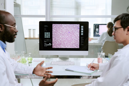 Middle aged Black man and young adult man discussing laboratory results in front of computer monitor displaying microscopic medical image, scientific equipment on tableの写真素材